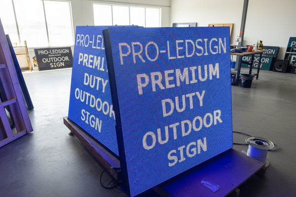 A rectangular outdoor LED sign with the text 'PRO-LEDSIGN' illuminated in blue, placed on a wooden surface in a dark environment.
