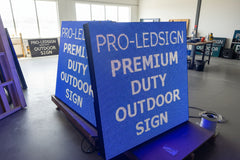 A rectangular outdoor LED sign with the text 'PRO-LEDSIGN' illuminated in blue, placed on a wooden surface in a dark environment.
