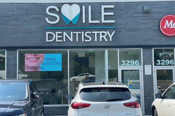 A digital LED sign on a store's window displaying the text 'SMILE DENTISTRY' with a heart symbol, surrounded by a brick wall and a parking lot with cars in front of the store.
